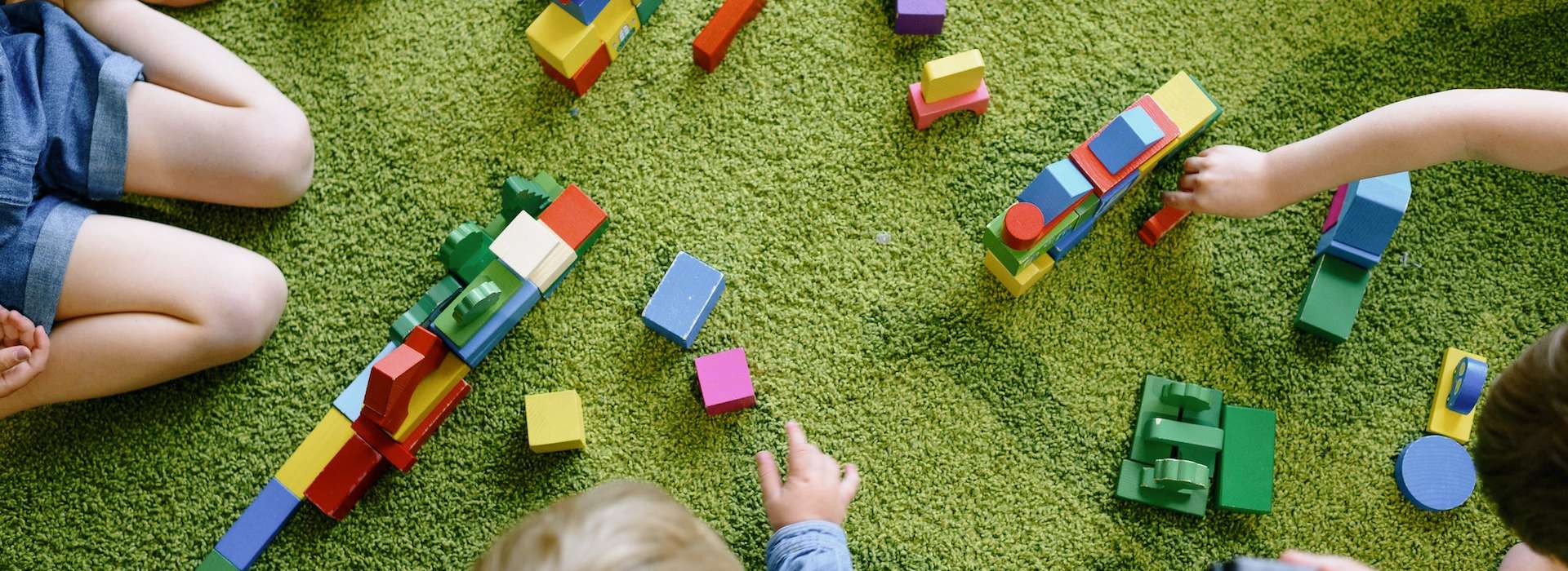 Kindertagesbetreuung. Das Symbolbild zeigt angedeutet drei Kinder mit Spielzeug auf einem grünen Teppich.